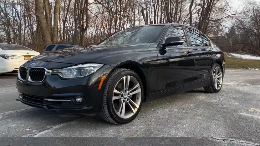 A Black BMW Sedan, Likely a 3 Series, Parked in An Outdoor Lot with Other Vehicles and Bare Trees in The Background, Showcasing Its Sleek Design and Performance-Oriented Stance