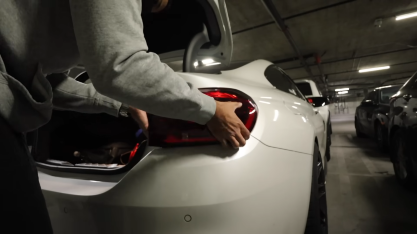 A Person Is Seen Working on The Taillight of A White Sports Car in A Dimly Lit Parking Garage, Seemingly Customizing or Repairing the Vehicle