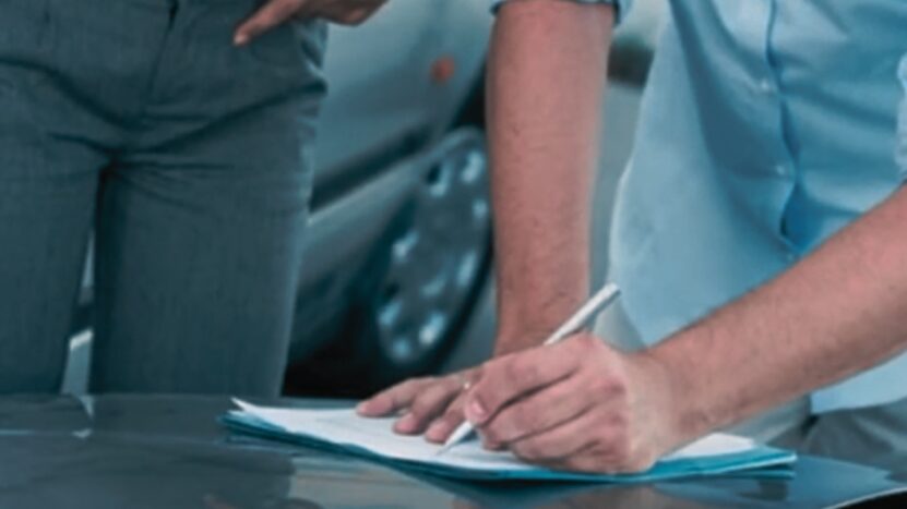 Two Individuals Are Exchanging Information After a Car Accident, with One Person Writing on A Document While the Other Stands Nearby